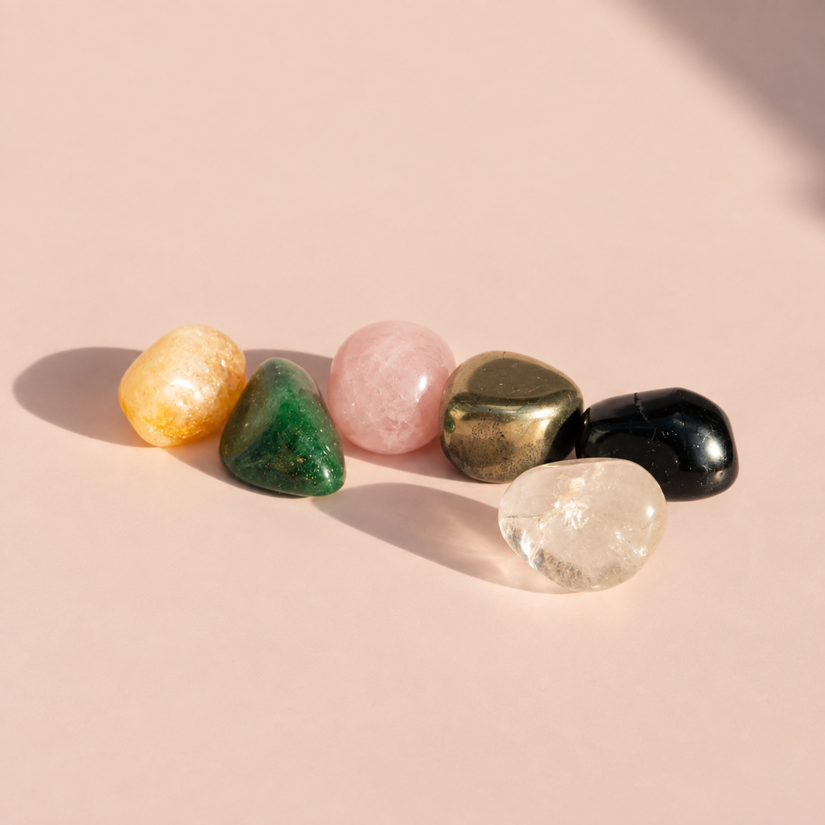 Six polished tumbled stones in Pyrite, Black Tourmaline, Clear Quartz, Citrine, Rose Quartz, and Green Aventurine arranged in a row on a soft pink background.