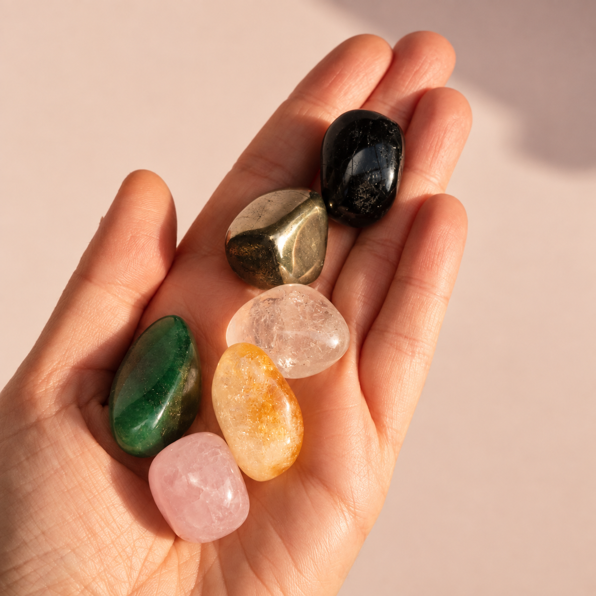 A close-up of a person's hand holding six polished tumbled crystals in Pyrite, Black Tourmaline, Clear Quartz, Citrine, Rose Quartz, and Green Aventurine, against a soft beige background.