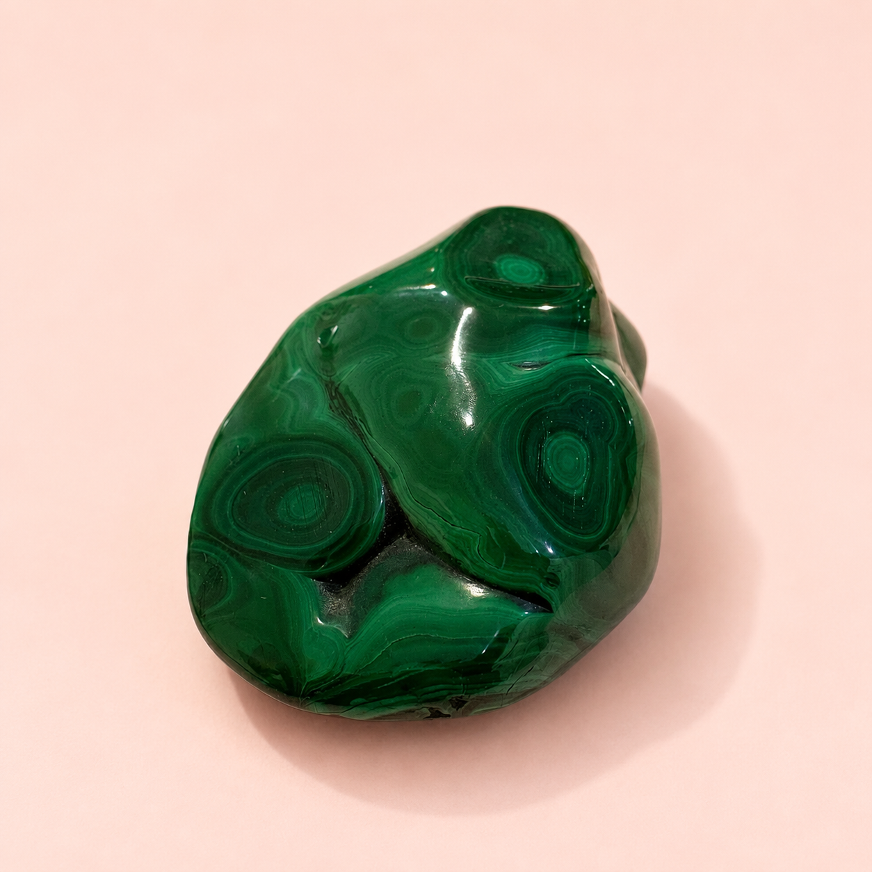 A polished green malachite stone with natural concentric band patterns, resting on a soft pink background.