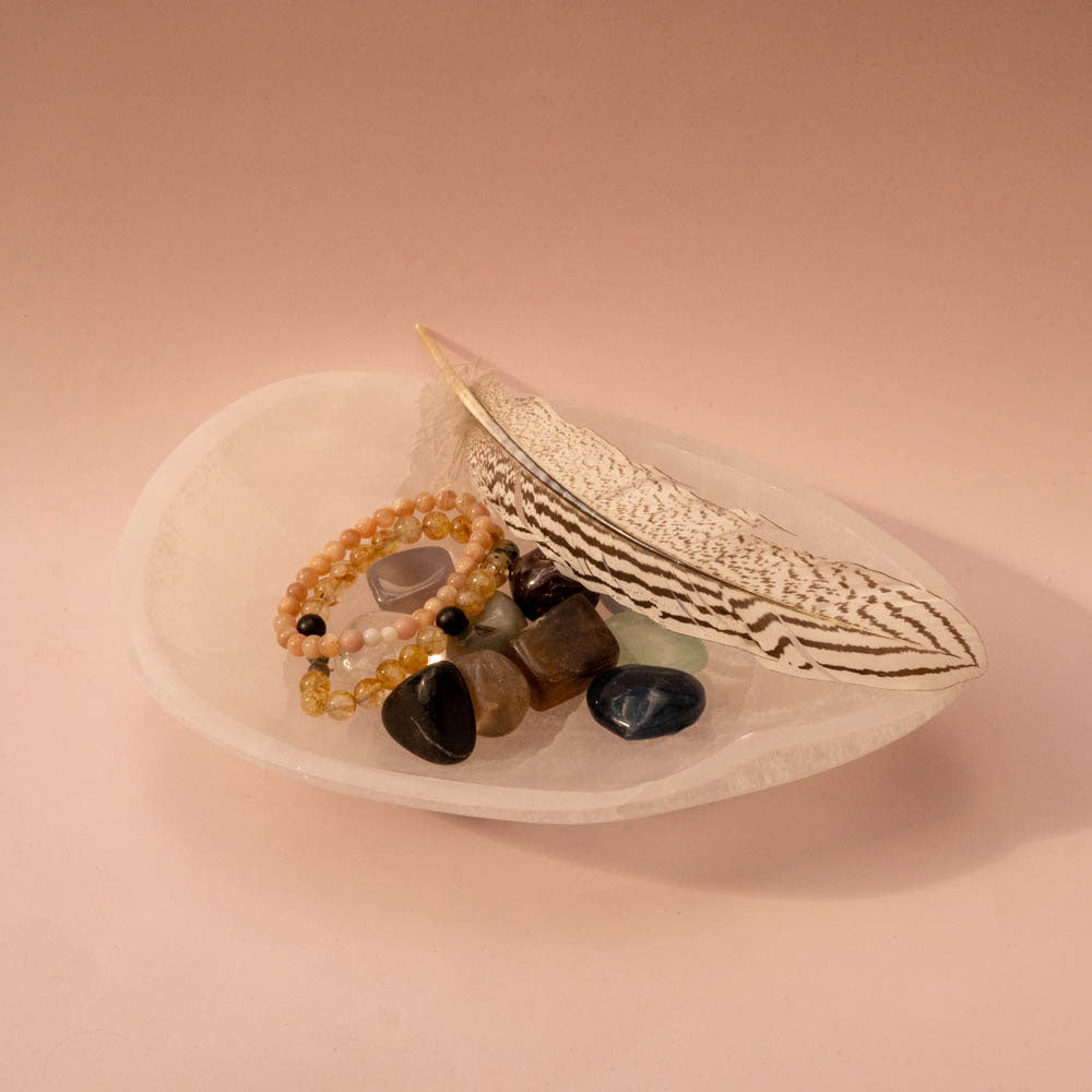 Selenite Crystal dish with a feather, stones, and beads on a pink background