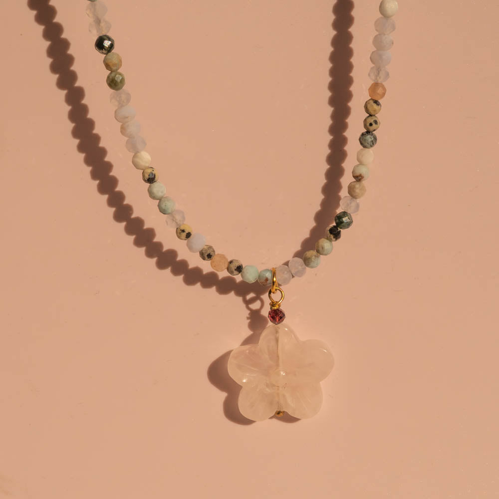 A beaded necklace with a pale pink rose quartz flower pendant, displayed on a warm peach-pink background. The necklace features multiple strands of faceted beads in various colors including green, white, and brown, with a small gold ring connector and a tiny red bead above the pendant.
