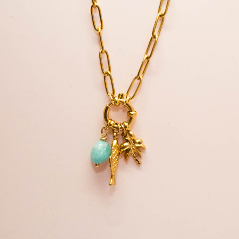 A gold paperclip chain necklace with a ring clasp, featuring three charms: a small gold fish, a gold palm tree, and a light blue oval bead, displayed on a light pink background.