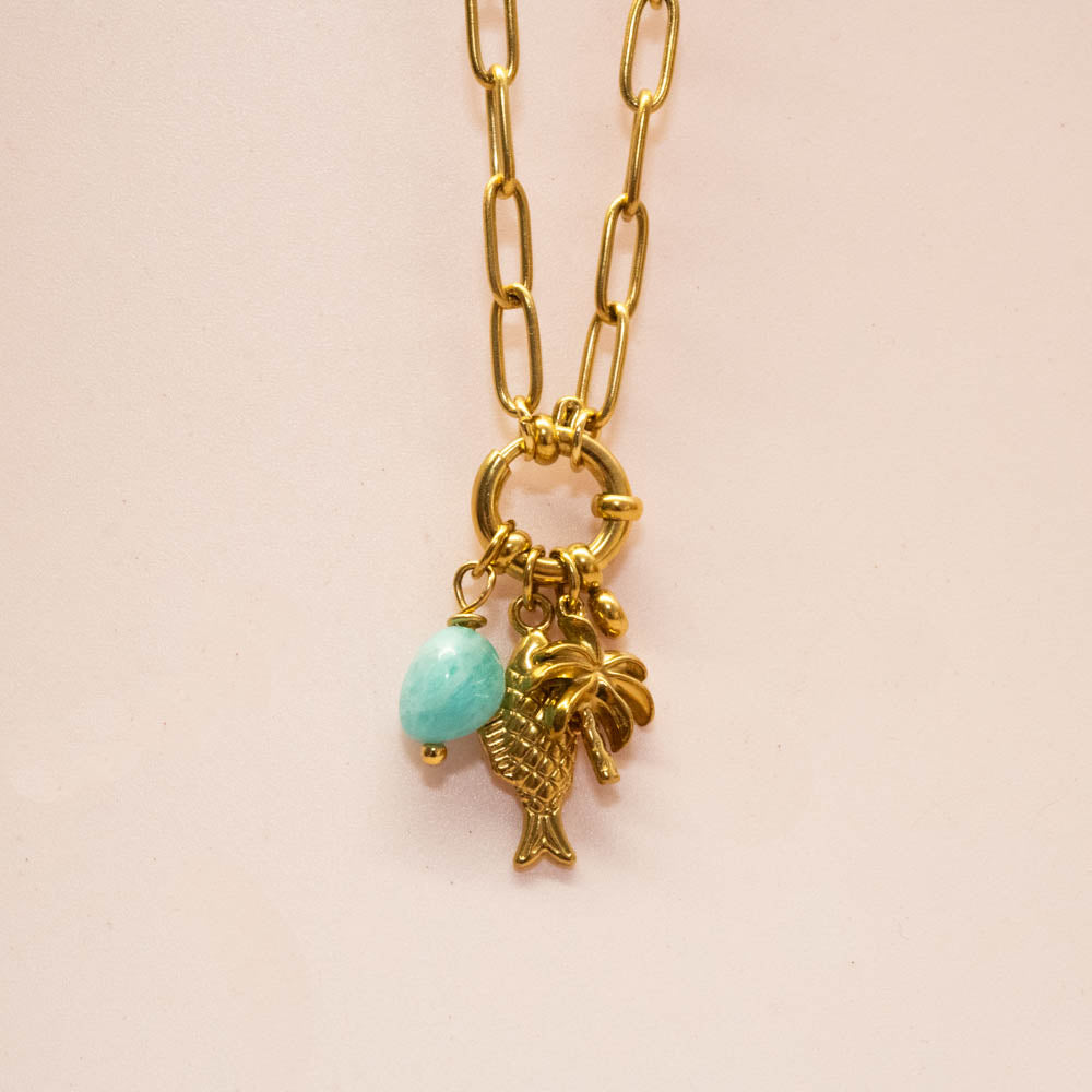 Gold paperclip chain necklace with a ring clasp holding a gold fish charm, a small gold palm tree charm, and a light blue oval bead charm, shown on a pale pink background.
