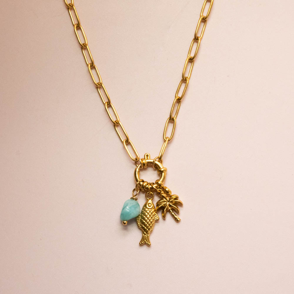 A gold paperclip chain necklace featuring a lobster clasp with a small gold fish charm, a palm tree charm, and a light blue bead charm, displayed on a soft pink background.