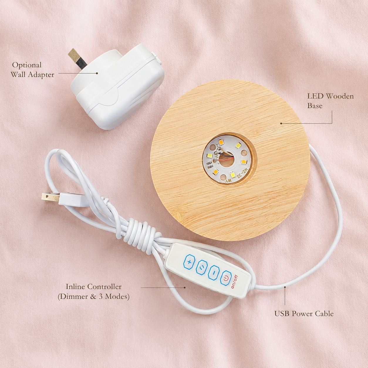 A round LED wooden base lamp with a light natural wood finish, shown with its white USB power cable, inline controller, and an optional white wall adapter on a soft pink fabric background. Text labels identify the components.