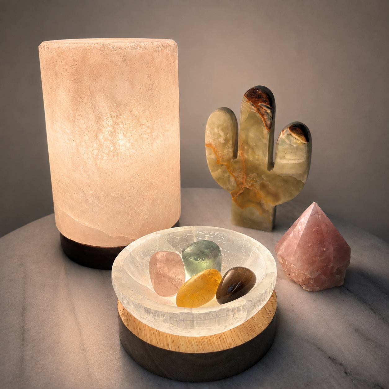 A glowing salt lamp stands on the left, while a clear crystal bowl with polished stones sits in front. A carved cactus and a pink crystal tower are placed behind on a gray surface against a neutral background.