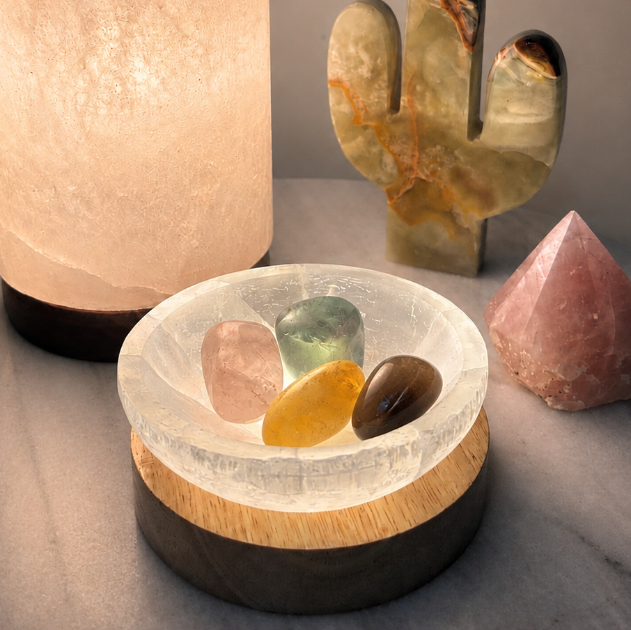 A clear crystal saucer with four polished tumbled stones sits on a dual-tone wooden base, styled on a marble surface with a glowing salt lamp, a cactus crystal, and a pink pyramid crystal in the background.